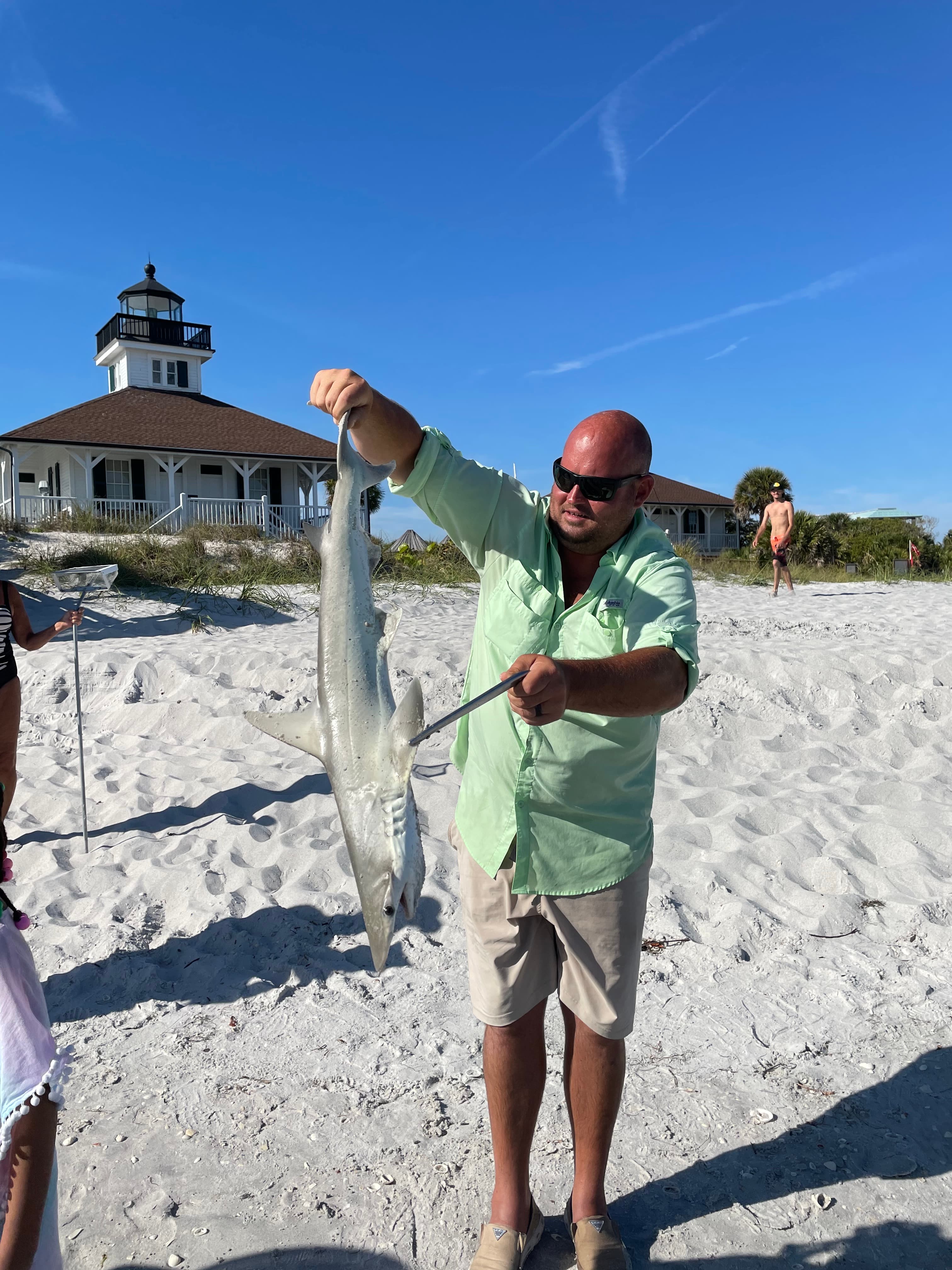 John on a white-sand beach holding a small shark he caught, lighthouse behind him